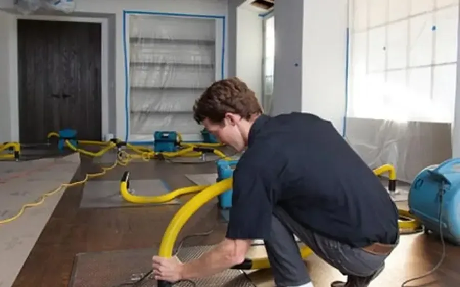 A technician connects drying hoses to floor mats beside blue equipment on hardwood
